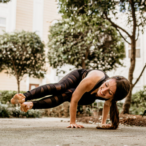 A woman with medium-length brunette hair in a pony tail and black athletic wear does an arm balance yoga pose on a sidewalk surrounded by trees