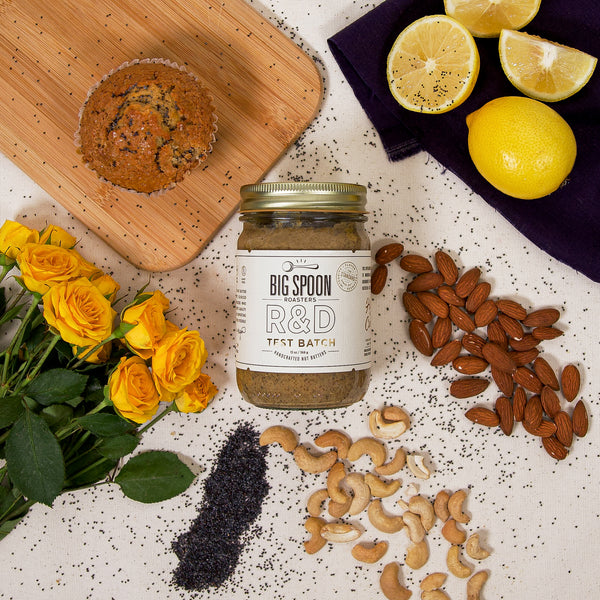 13oz Glass Jar of Lemon Poppy Seed Nut Butter with cashews, almonds, poppy seeds, lemons, and muffin, and yellow roses all lay on a white linen background