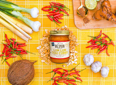 13oz glass jar of Lum Lum Thai Curry nut butter on golden fabric surrounded by chilis, lemongrass, coconut, peanuts, cashews, and shallots.