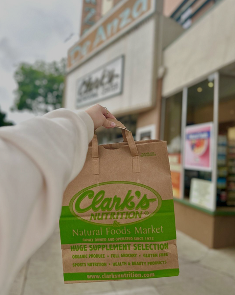 Girl's hand holding a brown paper bag that says Clark's Nutrition on it outside of a Clark's Market store