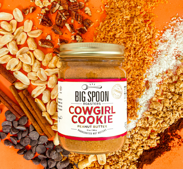 13oz glass jar of Cowgirl Cookie Peanut Butter with coconut flakes, pecan pieces, cashews, cinnamon sticks, chocolate chips, sea salt, granola, and vanilla powder spread out on an orange background.