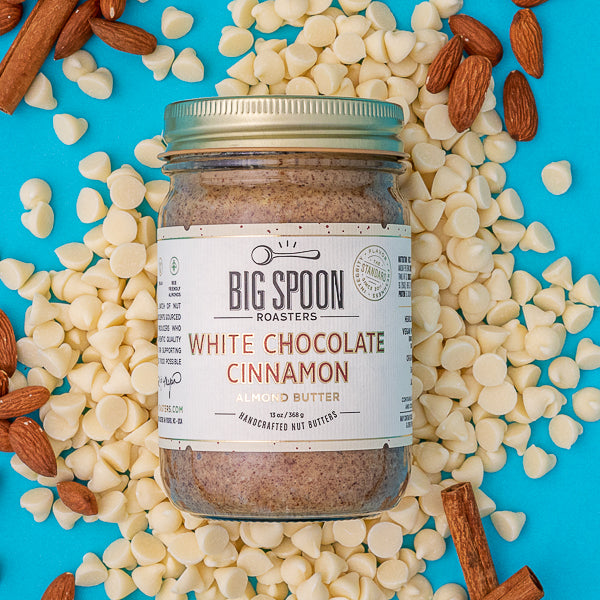 A jar of White Chocolate Cinnamon Almond Butter lays atop a bed of white chocolate chips, almonds, and cinnamon sticks.