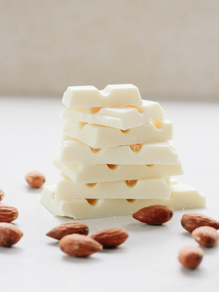 A stack of white chocolate stands vertically in front of a white background with almonds spread around it.