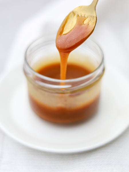 Caramel Sauce in a small glass jar with a gold spoon pulling some caramel out on top of a white saucer and white background.