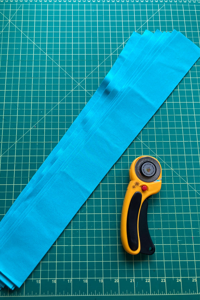 6 strips of binding fabric on a green cutting mat with a rotary cutter beside it.