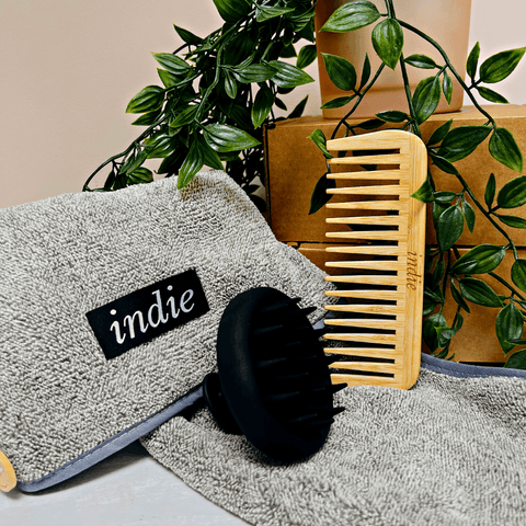 scalp massager, bamboo hair turban and bamboo comb laying on surface against box with plant