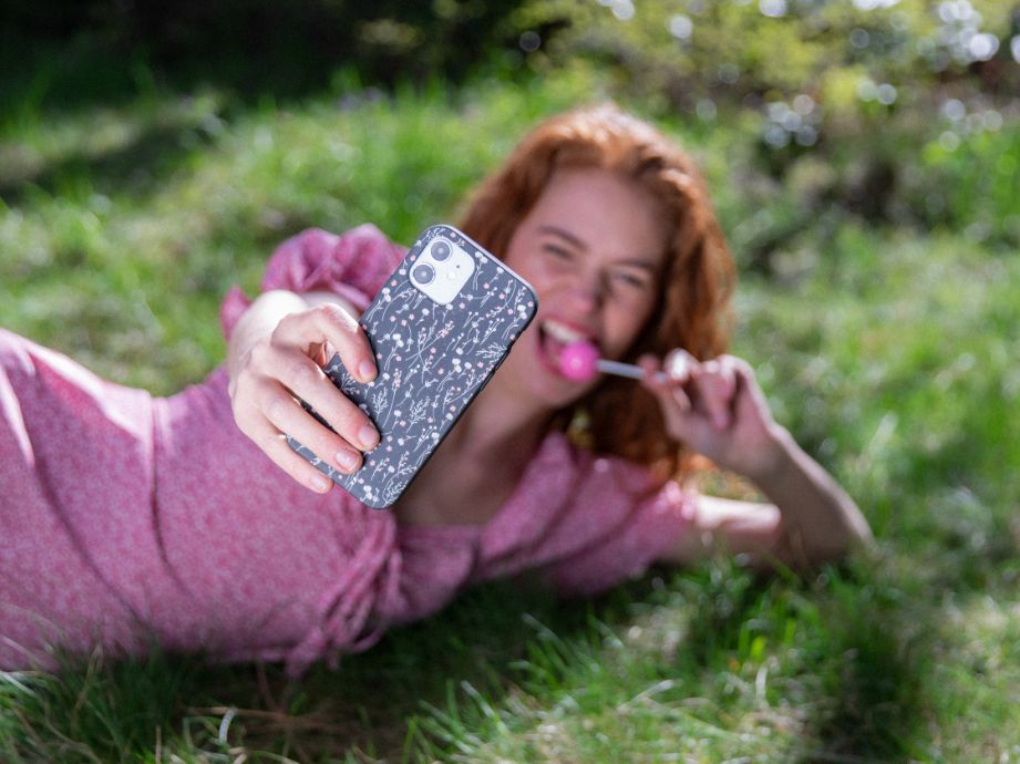 woman laying in a field with a lollipop and holding floral phone case towards camera