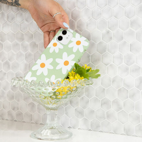 person sticking a phone case with white daisies on it into a glass with flowers