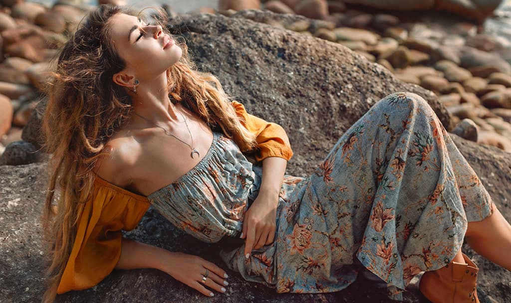 Mujer joven con un vestido bohemio sentada en una roca disfrutando del sol.