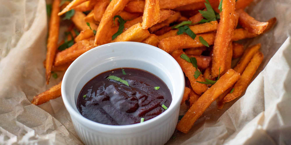 White ramekin filled with dark bbq sauce with green garnish on natural toned paper with orange sweet potato fries 
