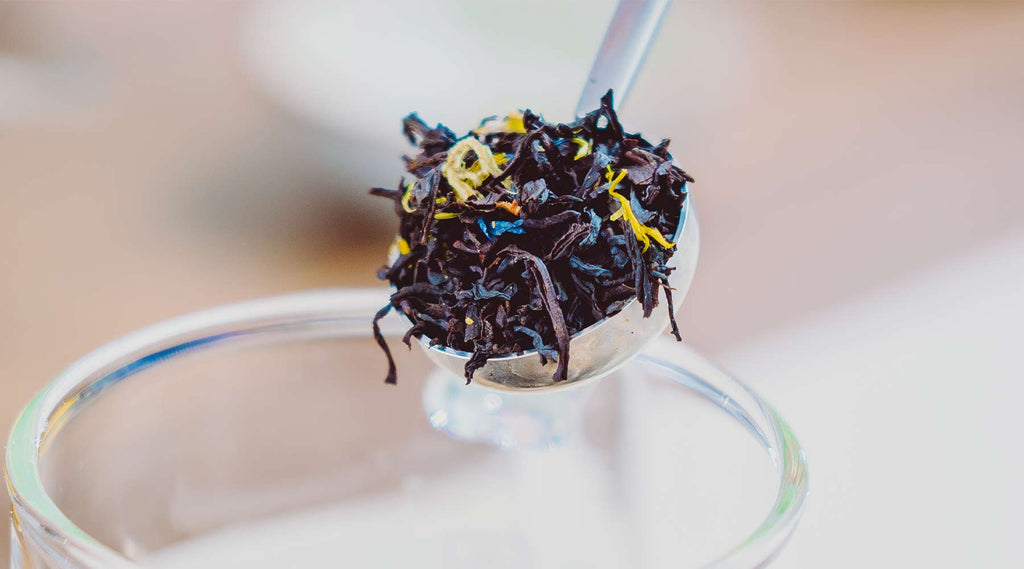 Metal Spoonful of black loose leaf tea leaves with blue corn flowers and yellow citrus rind throughout