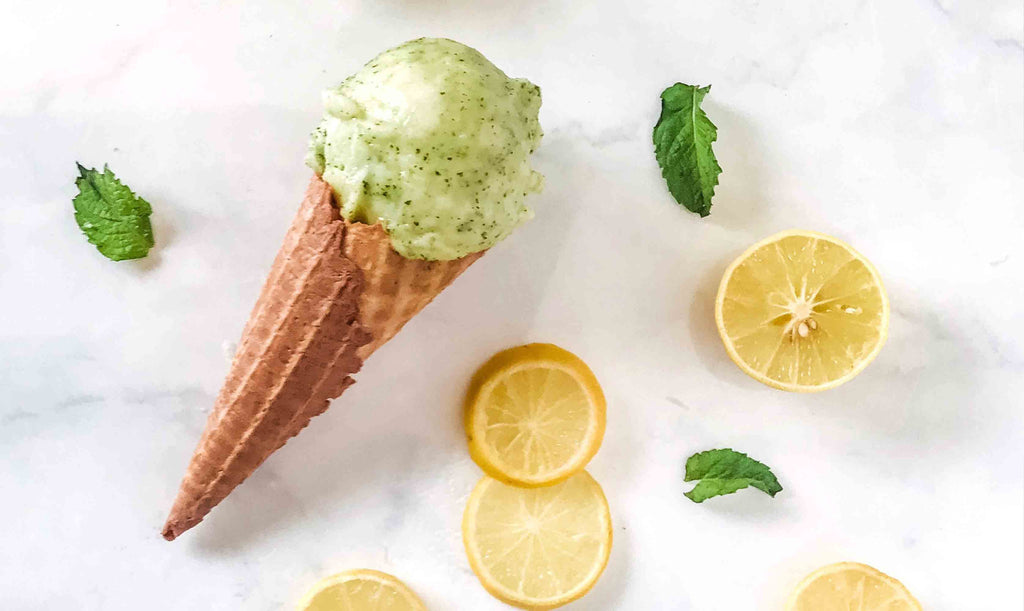 Waffle ice cream cone topped with green ice cream scoop on white marble table top, with scattered lemon slices and green leaves