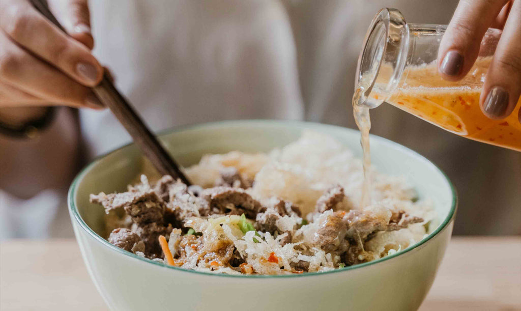 Bowl of rice with mixed vegetables and meats with dressing being poured over from glass jar and chop sticks in hand