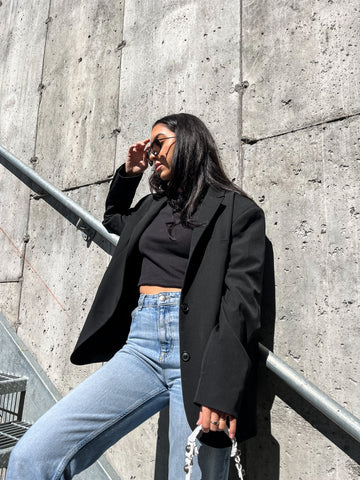 Fille dans un blazer noir avec un jean noir en denim devant un mur de béton avec des objets de créateurs