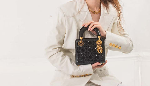 mini lady dior en cuir verni noir avec une quincaillerie dorée tenue par une fille en costume blanc avec un tour de cou dior doré