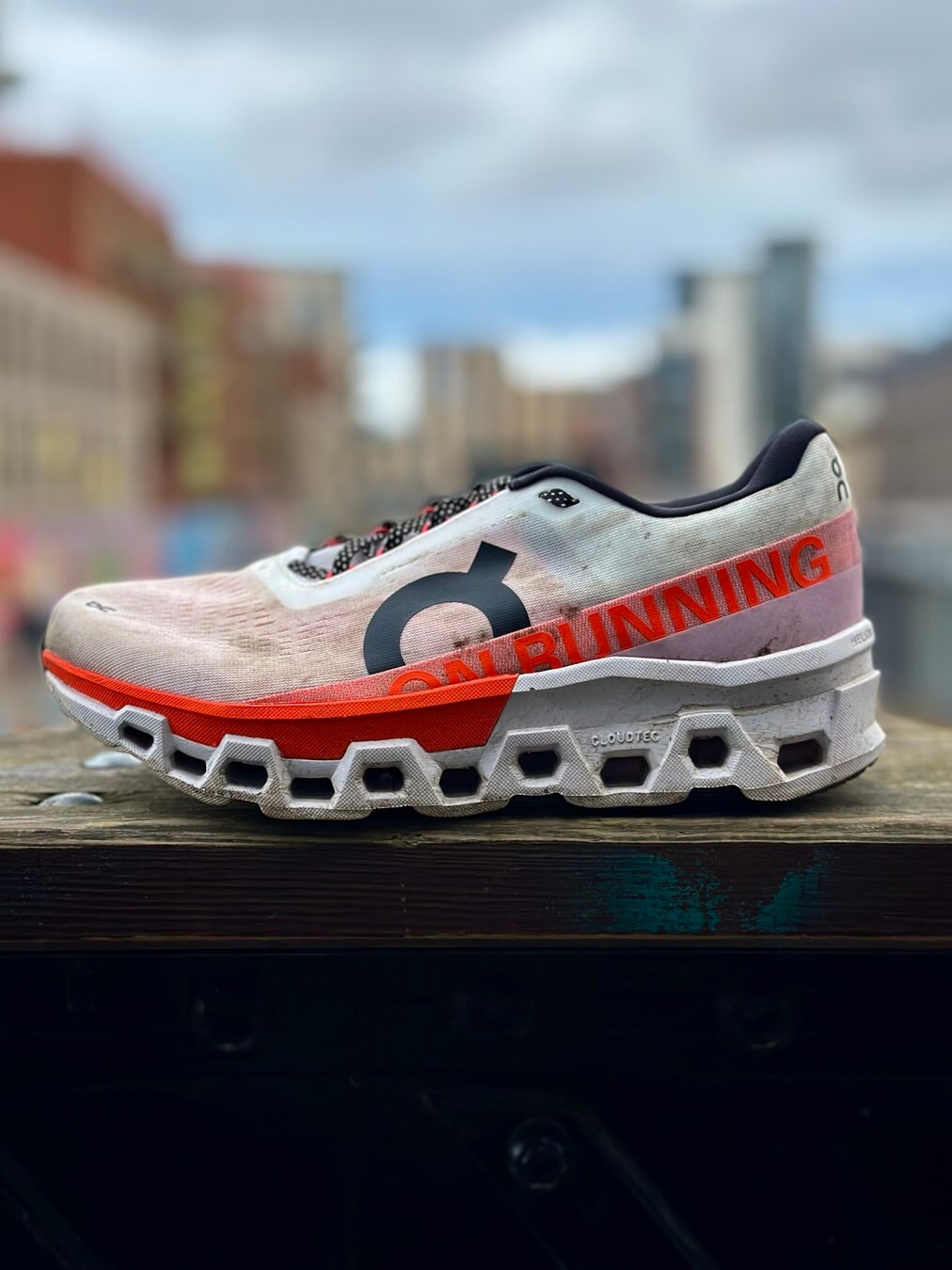 On Cloudmonster 2 running shoe in Undyed/Flame colourway perched on wooden railing with cityscape in background