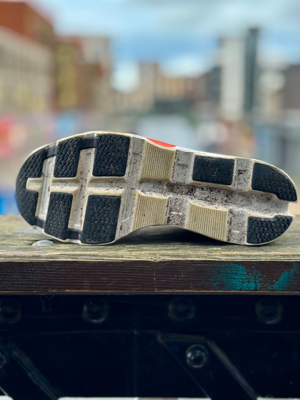 Outsole of On Cloudmonster 2 running shoe seen perched on timber rail with cityscape in background