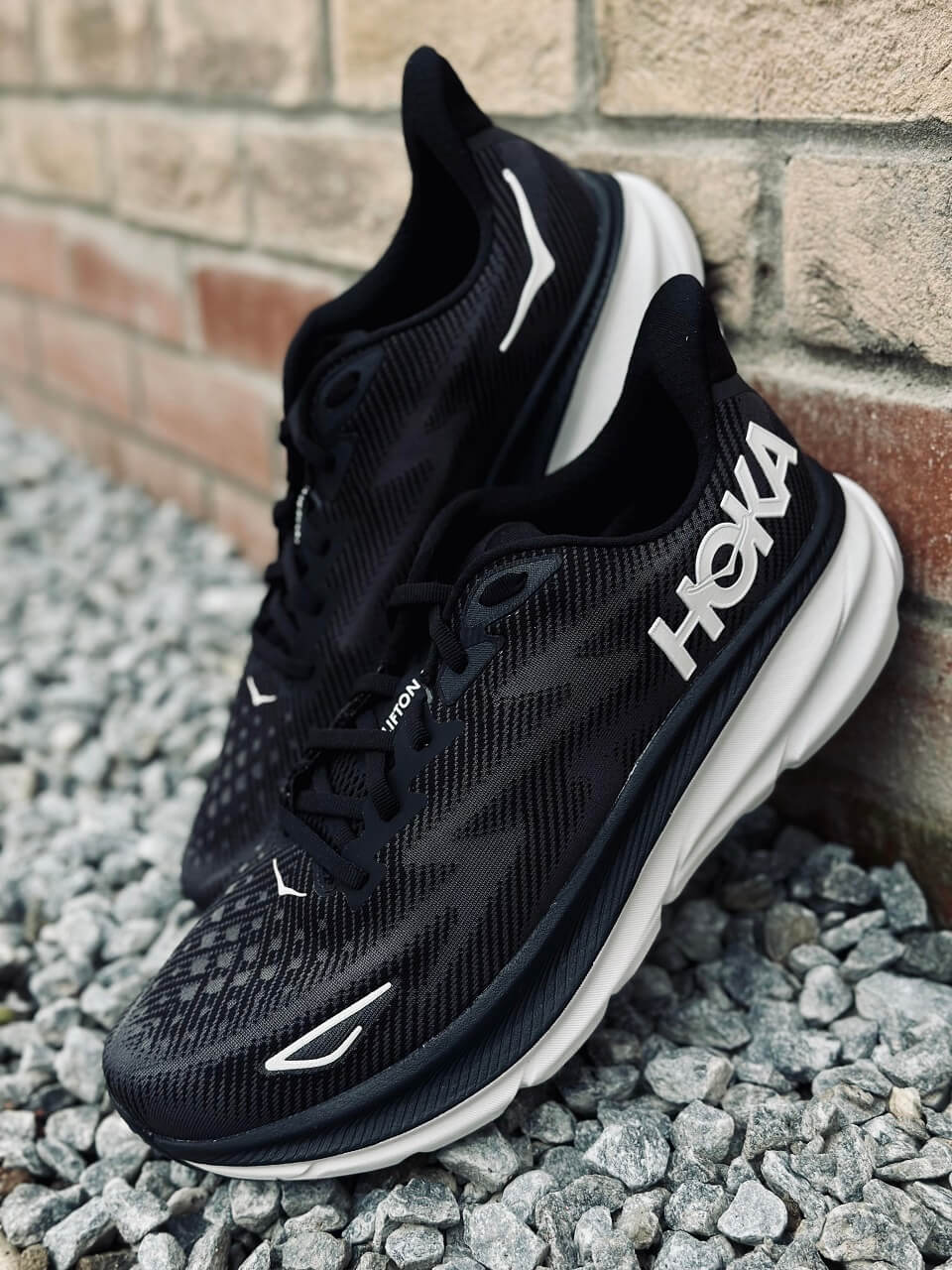 HOKA Clifton 9 running shoes in black propped up against a brick wall