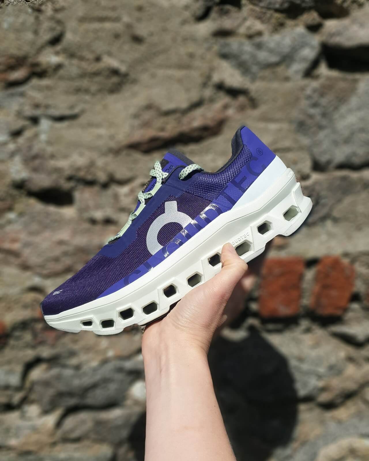 Hand holding an On Cloudmonster running shoe in purple against a stone wall