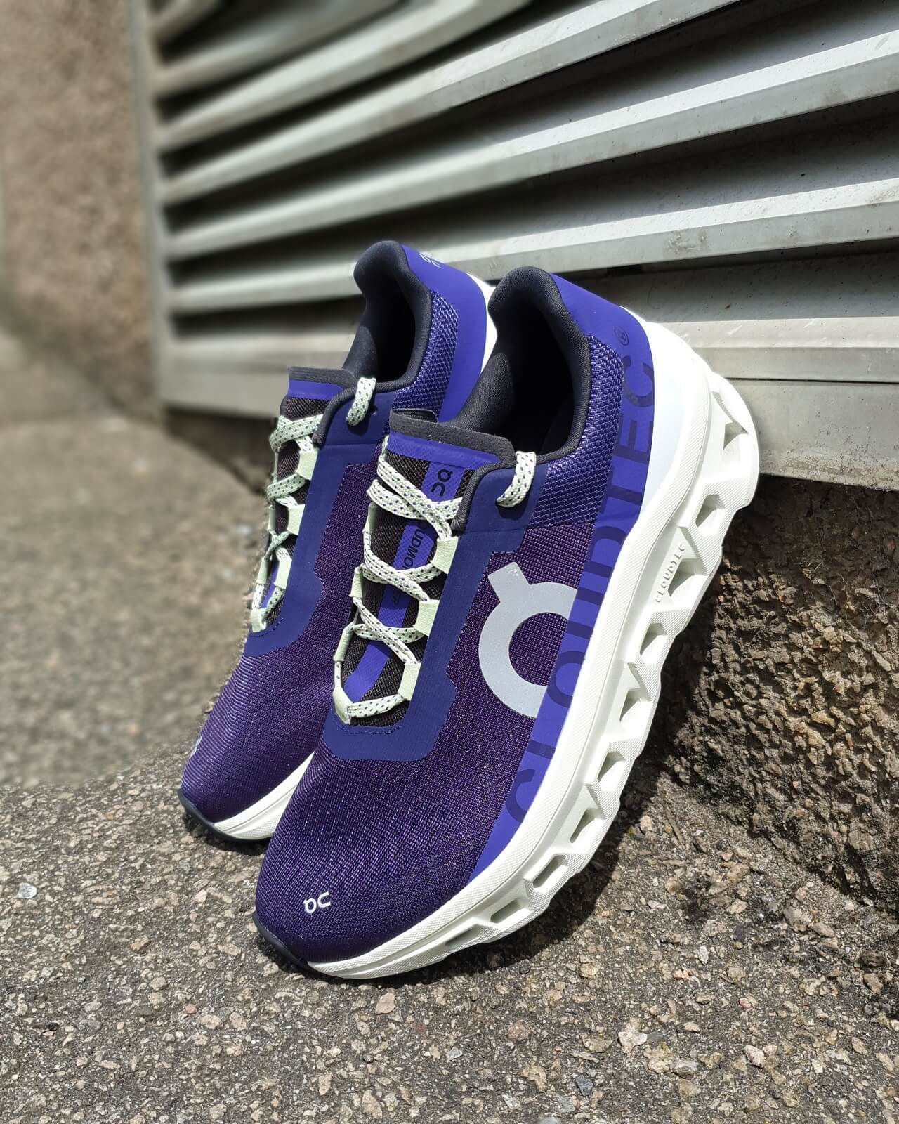 On Cloudmonster running shoes in purple perched against a wall with a ventilation grid behind