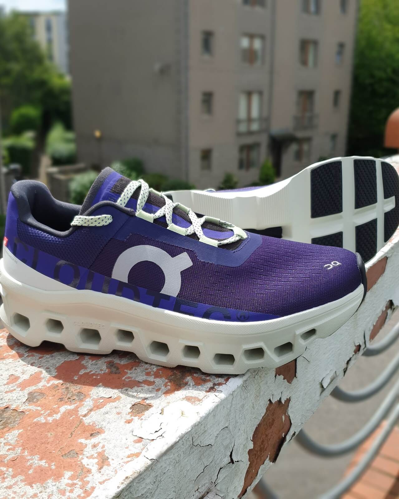 On Cloudmonster running shoes in purple perched on wall with urban tower blocks in the background