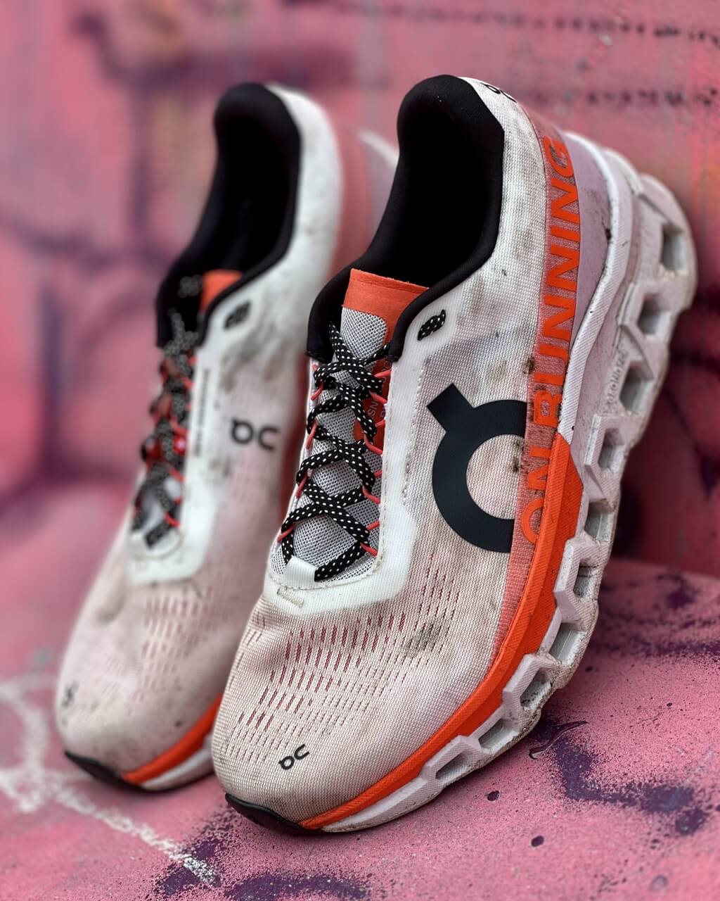 On Cloudmonster 2 running shoes in Undyed/Flame colourway propped against a pink painted wall