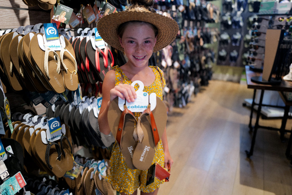 A little girl is smiling holding up a pair of Bethany Hamilton sandals.