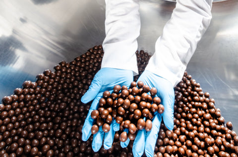 A panner wearing blue gloves holding perfectly coated panned chocolate, highlighting the result of skilled chocolate panning