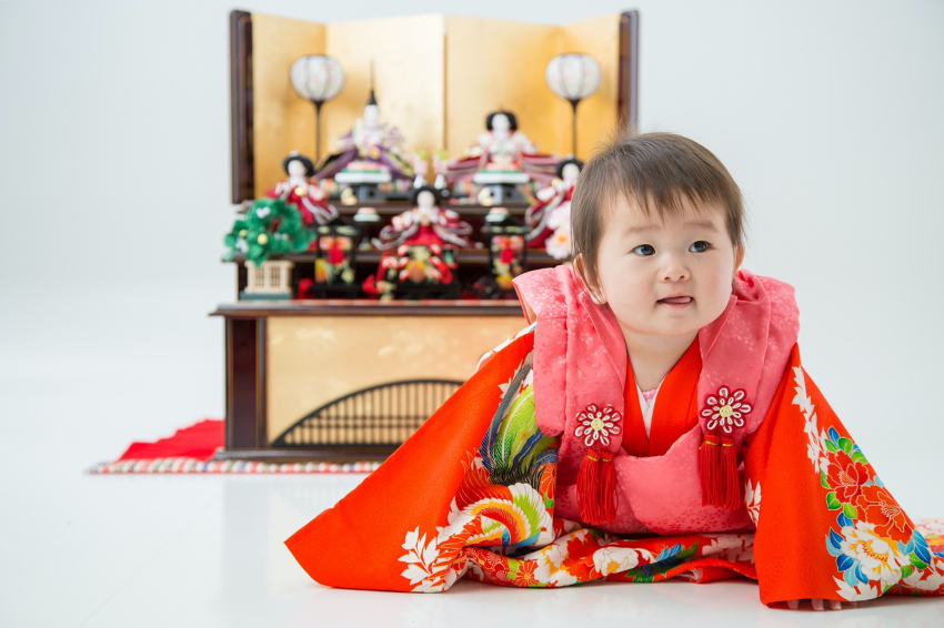 A baby girl in red kimono crowing infront of Hina dolls 