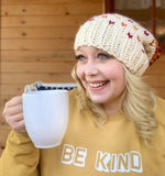 Suzanne holding a white mug dressed in a goldenrod "Be Kind" sweatshirt and knit beanie hat