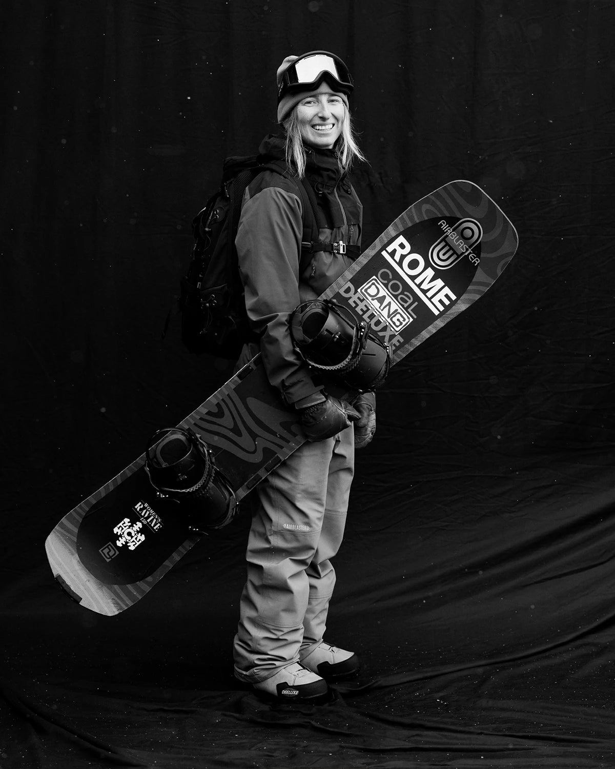 Monochrome image of a snowboarder holding a ROME snowboard featuring COAL DANG, DEELUXE bindings, and AIRBLASTER branding. The board's opposite side displays ROME RAVINE. The snowboarder wears DEELUXE boots and AIRBLASTER pants.