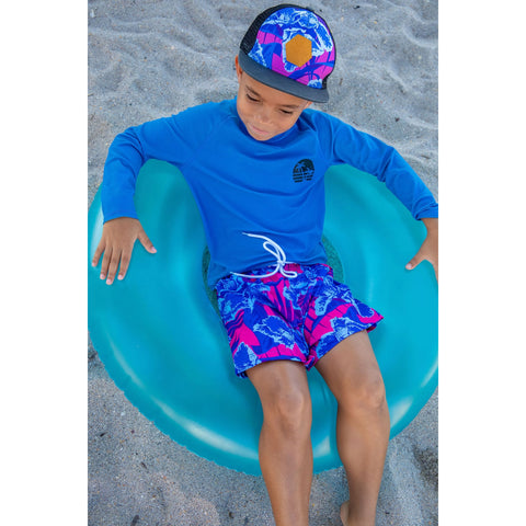 boy laying on the beach sand in a trendy boys rashguard and floral trendy boys swimwear from Blueberry Bay Swimwear for kids