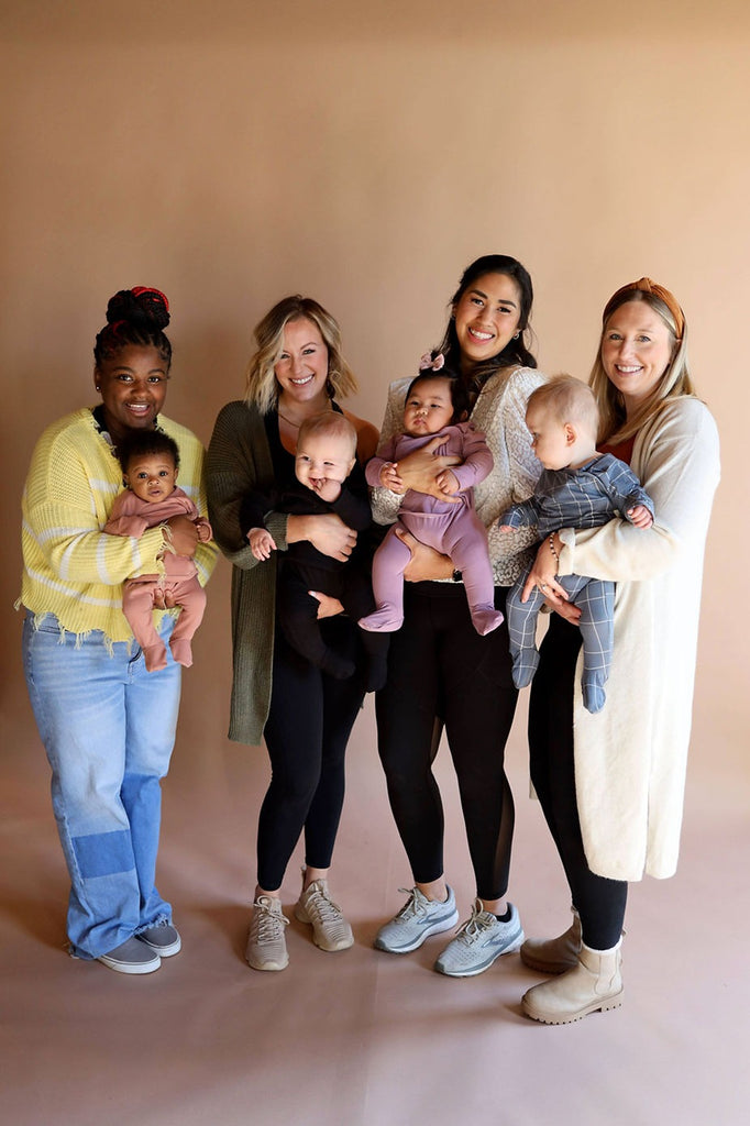 Photo of four moms posing with their babies in front of a neutral background. The babies are wearing Bonsie Skin to Skin Footies.