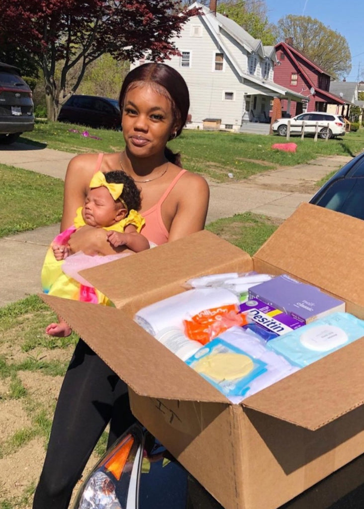 A woman holding her baby stands behind Bonsie’s Welcome Box, filled with food, toiletries, and a book. The background shows houses under a clear sky.