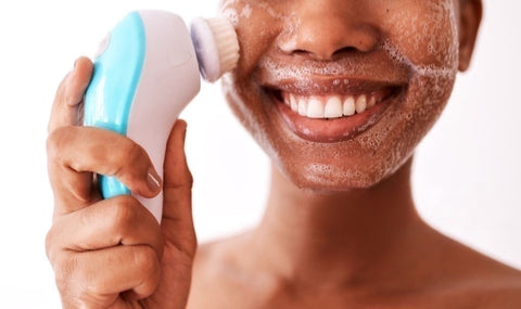 alt="Young african american girl double cleansing her face and creating suds with a blue and white electric face brush."