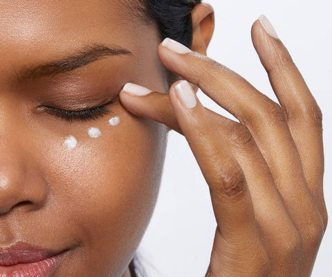 alt="Caramel complexion black woman applying three dots of eye cream under her left eye."