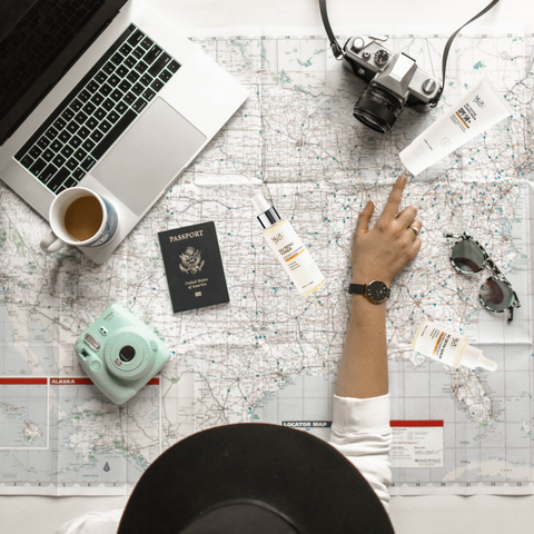 alt="World traveler with white shirt and black hat looking at a map with Macbook, a cup of coffee, cameras, passport, and NU2U Skincare products on map. Pointing at a tube of NU Shyne Sunscreen."