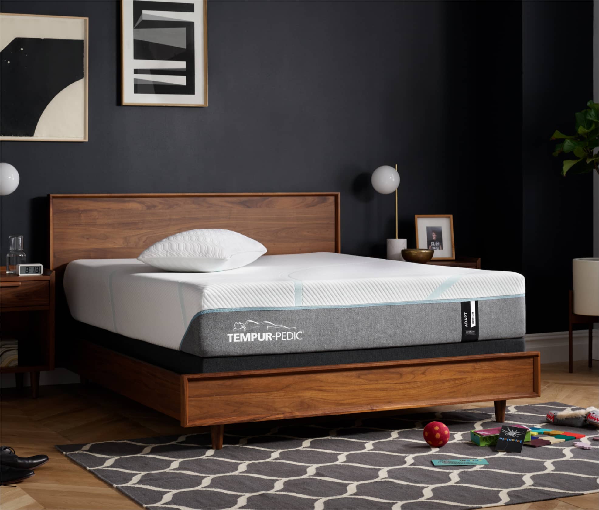 A Tempur-Pedic Mattress is set on top of a wooden bed frame that is against a dark blue wall. There are two black and white art pieces hung on the wall above it and a patterned gray and white rug beneath it.