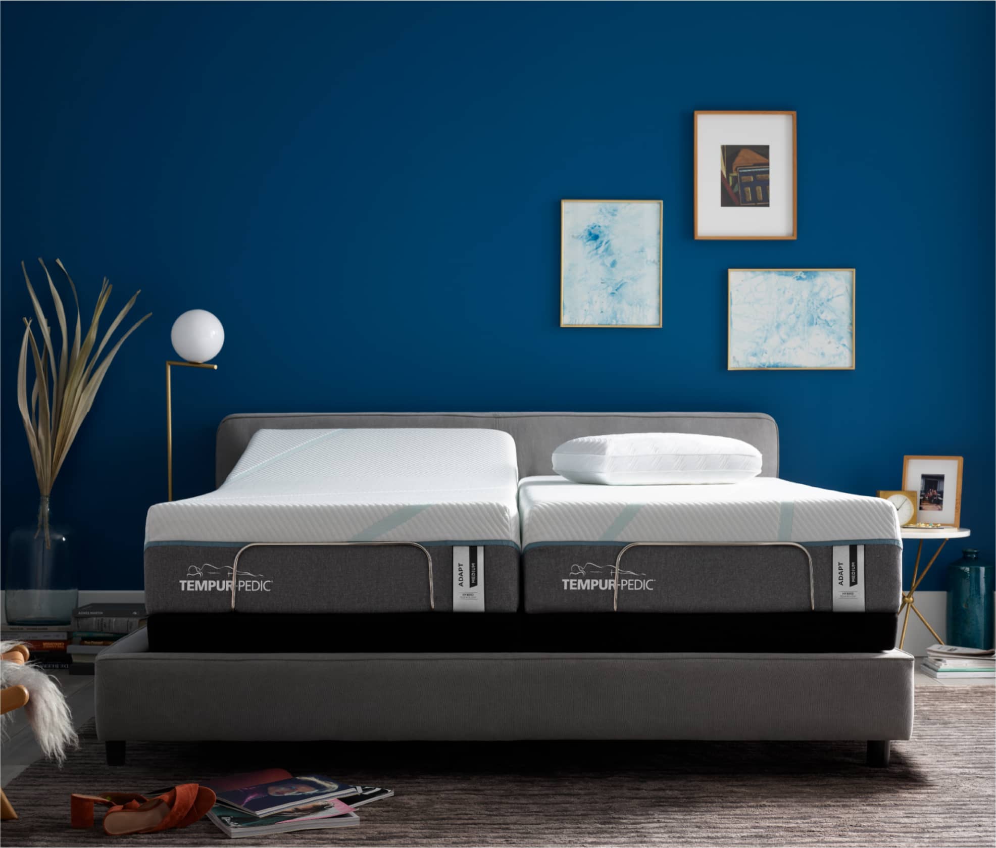 Two Tempur-Pedic Twin Mattresses sit on a king size gray upholstered bed frame, one of the mattresses is slightly elevated showing that the two mattresses can elevate independently. The bed is against a dark blue wall with three pieces of art work hung on the wall toward the right side of the bed.