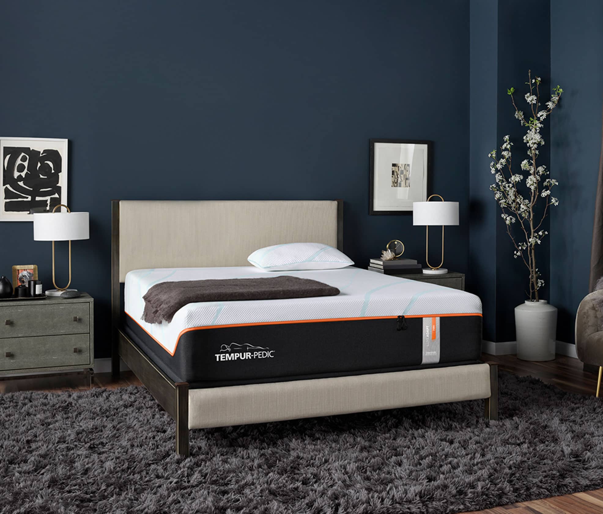 A Luxe Tempur-Pedic Mattress sits on top of a cream upholstered bedframe that is against a dark blue wall. There are two black and white artworks on either side hung on the wall and two gray wood nightstands.