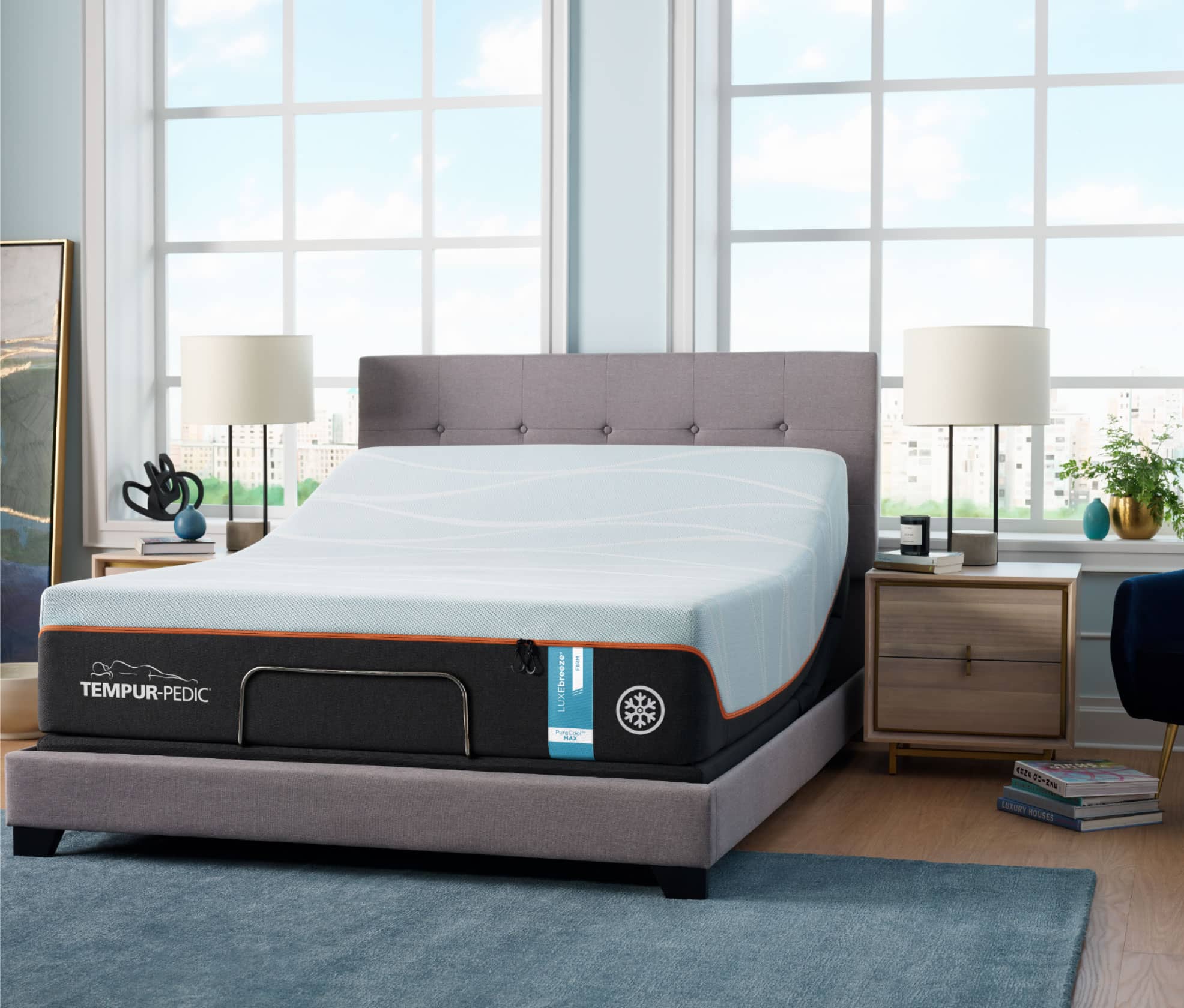 A LuxeBreeze Tempur-Pedic Mattress sits on a gray upholstered adjustable bed frame, the head of the mattress is slightly elevated. The bed sits against two large bright windows showing a clear blue sky outside.