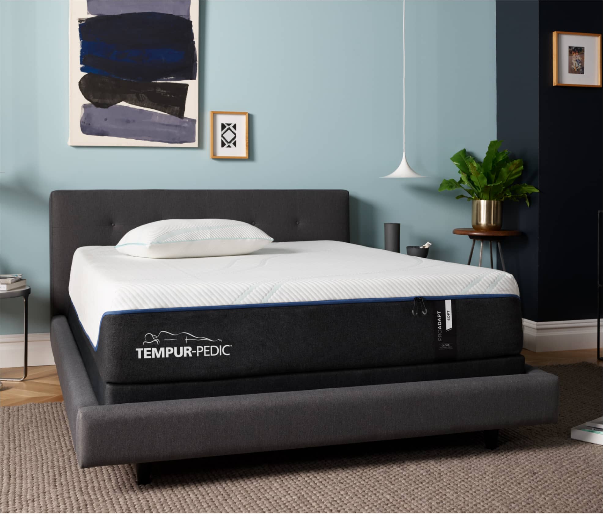 A Tempur-Pedic Mattress sits on top of a dark gray upholstered bedframe against a light blue wall. There is an abstract art of different shades of blue hung on the wall above it