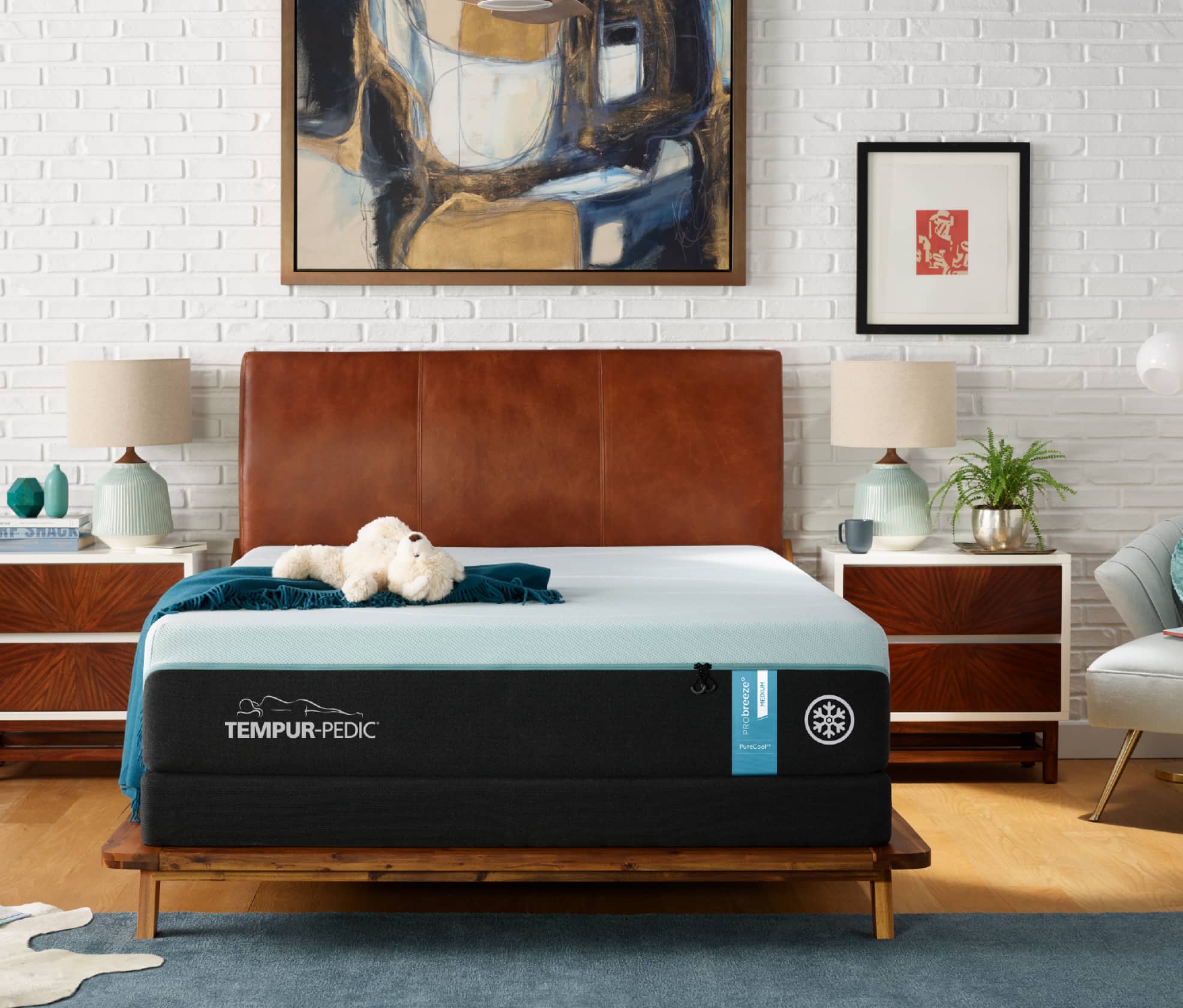 A ProBreeze Tempur-Pedic Mattress sits on a wooden bedframe with a brown leather headboard against a white brick wall. There is a gold, blue, and white abstract painting on the wall above the bed and two brown and white nightstands on either side of the bed.