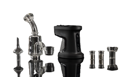 Alt text: "An assortment of dab rig equipment displayed on a reflective surface against a black background. On the left, there is a traditional glass dab rig with a transparent, curved neck and decorative glass elements. In the center, a modern black electric dab rig with a textured grip and digital display panel branded 'daab' stands prominently. To the right, various transparent glass components, possibly for customization, are neatly lined up, including different sized glass bowls and adapters, reflecting on the surface below.