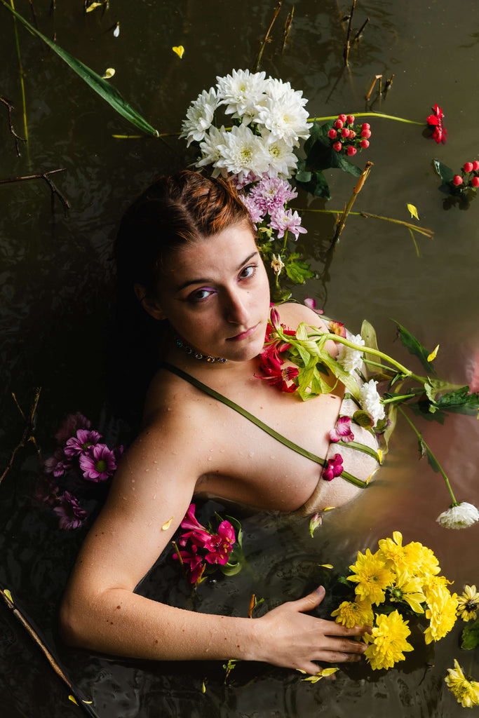 Interview with Rosie Lugg @rambosphotos. A girl is half submerged in murky water, surrounded by bright flowers. She looks up at the camera.