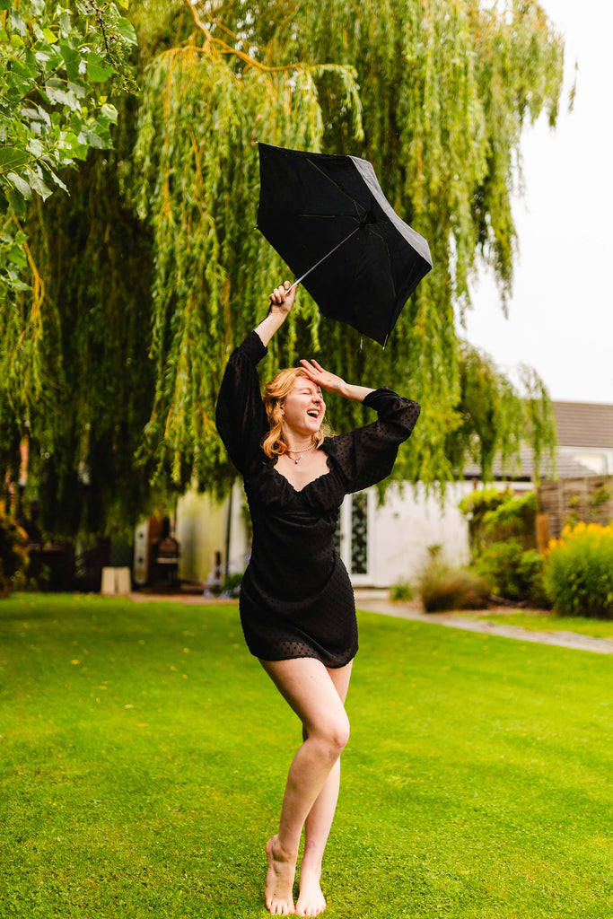 Interview with Rosie Lugg @rambosphotos. A girl in a black dress poses in the rain with a black umbrella. She looks happy.