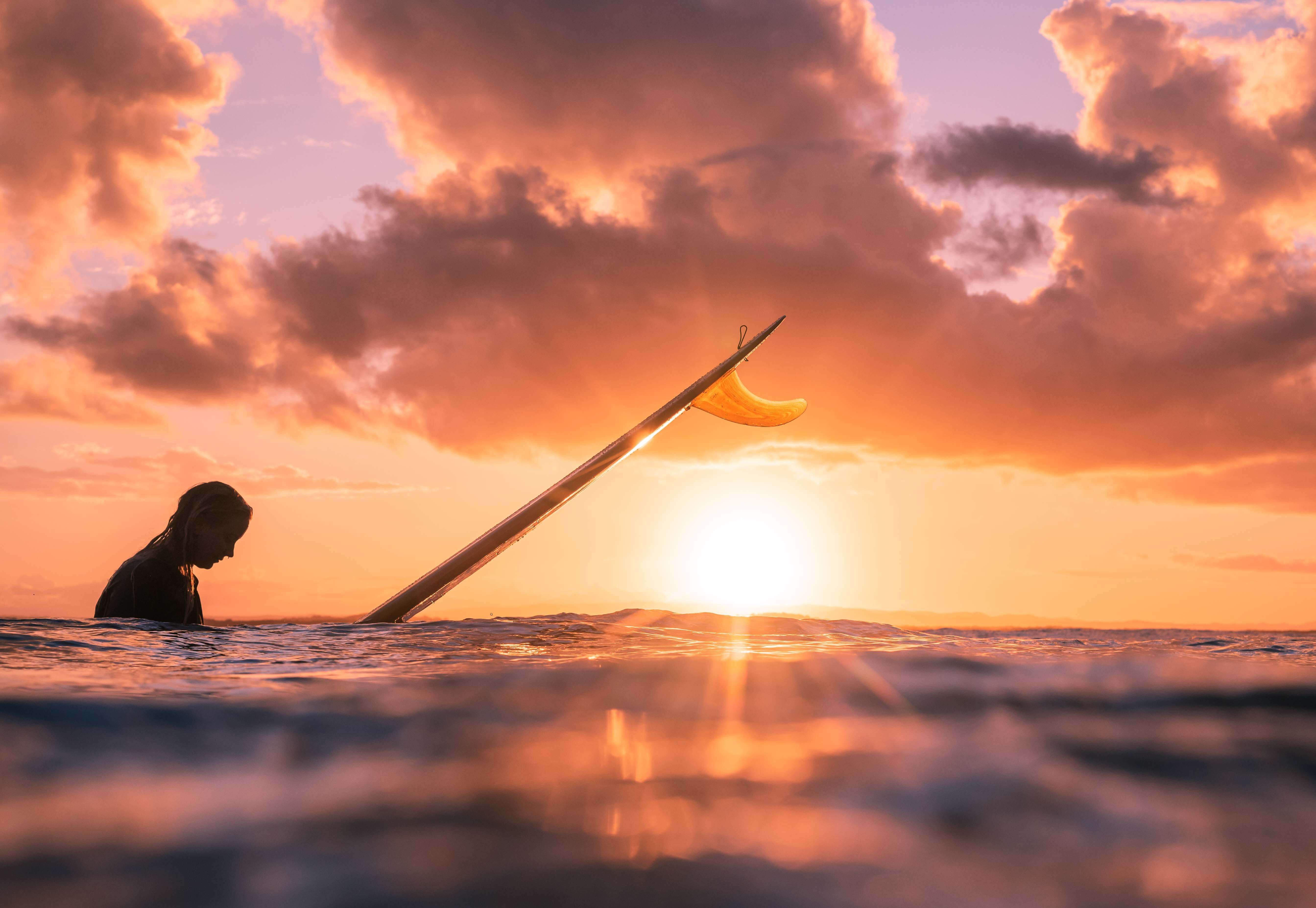 Sun sets over the ocean, lighting the sky up in orange hues as a surfer paddles past. Taken by photographer Hannah Prewitt