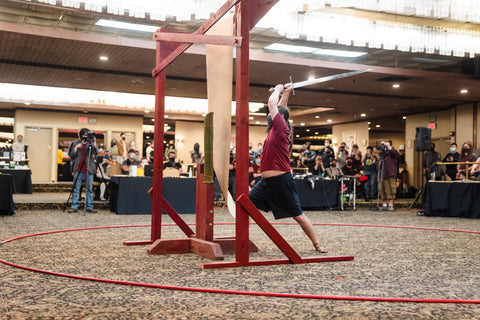 Competitor preparing to cut through paper hanging from red wooden stand while crowd watches at SoCal Swordfight 2022