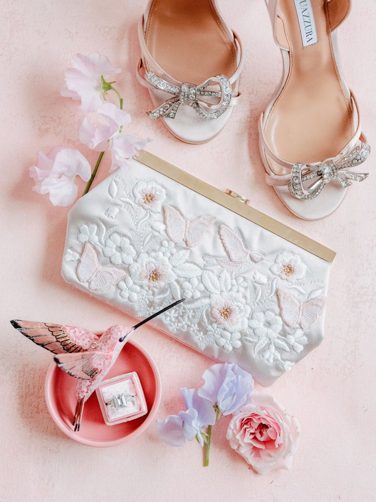 Custom handbag with white floral embroidery and pink butterflies and hummingbirds against a pink backdrop, styled with flowers, shoes, and a bridal ring