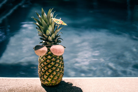 a pineapple placed by pool with sunglasses on and wearing cocktail umbrella